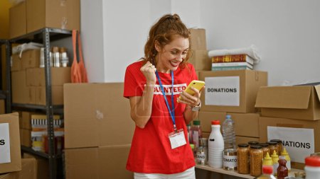 Photo for A cheerful young woman wearing a volunteer shirt enjoys a break with her phone in a donation center storeroom. - Royalty Free Image