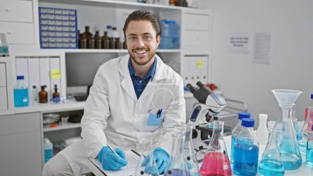 Photo for Young hispanic man scientist writing report smiling at laboratory - Royalty Free Image
