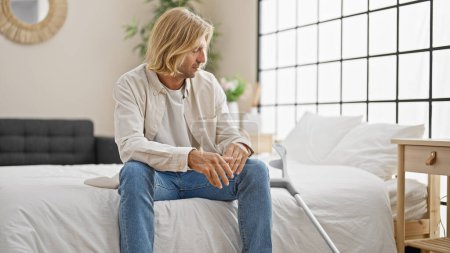 Téléchargez les photos : Un jeune homme aux longs cheveux blonds s'assoit pensivement sur un lit à l'intérieur, béquilles penchées à proximité. - en image libre de droit