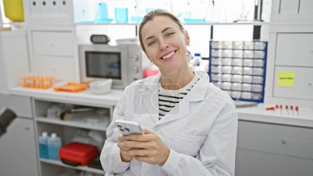 Foto de Mujer sonriente con bata de laboratorio usando smartphone en un entorno de laboratorio moderno - Imagen libre de derechos
