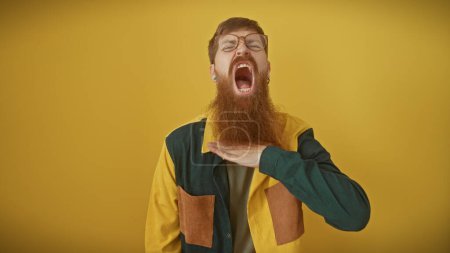 Photo for A bearded young redhead man in a colorful jacket makes a throat-cutting gesture against a yellow background, displaying anger or frustration. - Royalty Free Image
