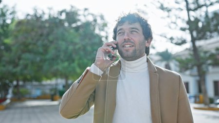 Homme hispanique vêtu occasionnellement parle au téléphone, rayonnant, au milieu des paysages verts du parc sous un ciel clair.