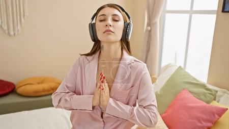 Eine ruhige junge Frau in rosa Pyjama meditiert auf einem Bett mit Kopfhörern in einer gemütlichen Schlafzimmeratmosphäre.