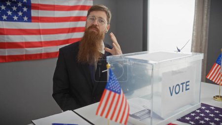 Téléchargez les photos : Un homme barbu fait un geste passionné près d'une urne électorale américaine, flanqué de drapeaux américains dans un cadre intérieur. - en image libre de droit