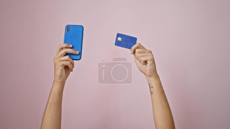 Brazos tatuados del hombre sosteniendo el teléfono inteligente y la tarjeta de crédito contra la pared rosa, que representa el pago en línea o compras.