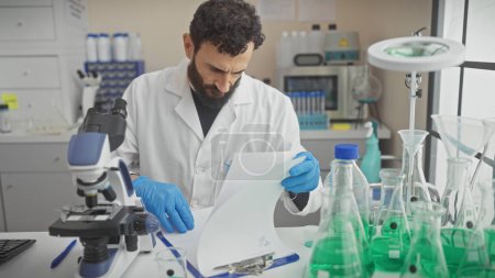 Foto de Un hombre barbudo con una bata de laboratorio examina un documento en un moderno laboratorio lleno de cristalería y un microscopio. - Imagen libre de derechos