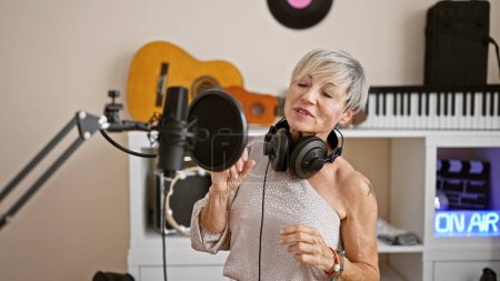 Reife Frau singt in einem Musikstudio mit Mikrofon und Kopfhörer und zeigt eine leidenschaftliche Performance in Innenräumen.