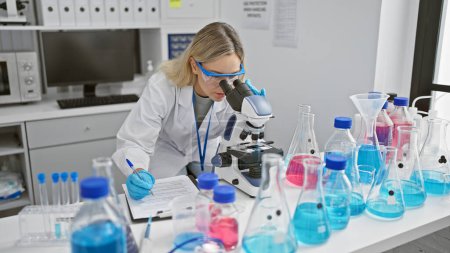 Téléchargez les photos : Une jeune scientifique prend des notes en utilisant un microscope dans un laboratoire rempli de verrerie et de produits chimiques. - en image libre de droit
