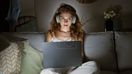 Photo for A young woman in casual clothing uses a laptop while wearing headphones, sitting on a couch in a cozy living room at night. - Royalty Free Image