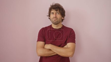 A skeptical young hispanic man with a beard is standing arms crossed against a pink wall, conveying a sense of contemplation.