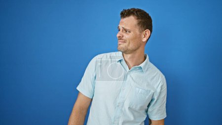 Selbstbewusster junger Mann mit lässigem Look und Bart posiert draußen vor leuchtend blauem Hintergrund.