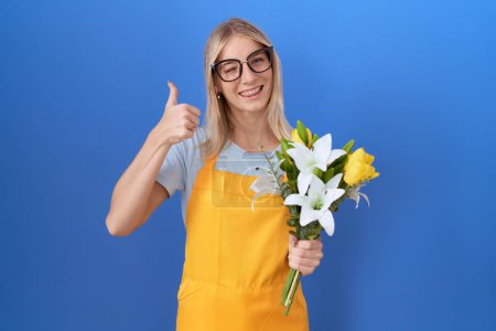 Téléchargez les photos : Jeune femme caucasienne portant un tablier de fleuriste tenant des fleurs faisant des pouces heureux geste avec la main. approuvant l'expression regardant la caméra montrant le succès. - en image libre de droit