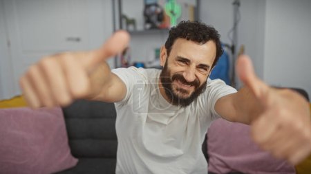 Téléchargez les photos : Un homme souriant avec une barbe donnant pouces vers le haut dans un salon confortable. - en image libre de droit