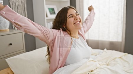 Eine fröhliche junge Frau dehnt sich, während sie auf einem Bett sitzt, und verkörpert Komfort und Glück in einem hellen Schlafzimmerinnenraum.