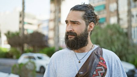 Ein bärtiger junger Mann mit trendiger Frisur steht lässig auf einer Straße in der Stadt und verkörpert urbanen Stil und entspanntes Selbstbewusstsein.