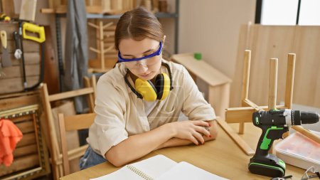 Photo for Focused woman wearing safety goggles and ear protection planning furniture construction in a woodworking workshop. - Royalty Free Image