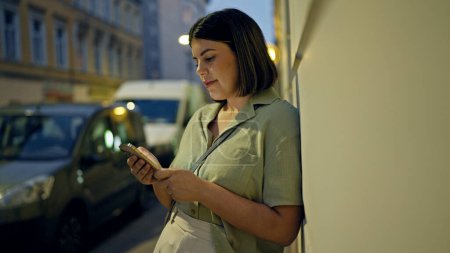 Photo for Young beautiful hispanic woman smiling happy using smartphone in the streets at night - Royalty Free Image