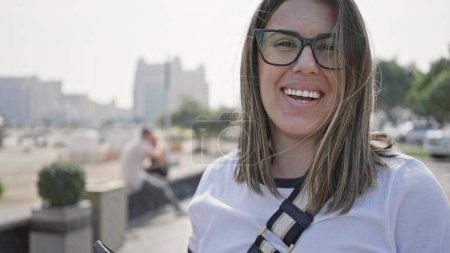 Eine lächelnde junge erwachsene Frau mit Brille, wie eine Brünette gestylt, genießt ein sonniges Stadtbild am Meer in Doha, Katar.