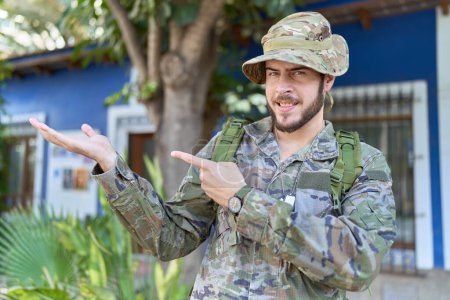 Photo for Young hispanic man wearing camouflage army uniform outdoors amazed and smiling to the camera while presenting with hand and pointing with finger. - Royalty Free Image
