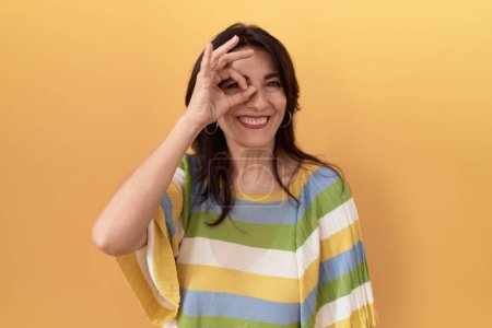 Foto de Mujer hispana de mediana edad de pie sobre fondo amarillo haciendo buen gesto con la mano sonriente, los ojos mirando a través de los dedos con cara feliz. - Imagen libre de derechos