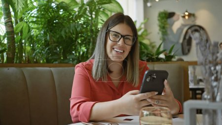 Téléchargez les photos : Femme souriante utilisant un smartphone dans un café avec des plantes et du verre d'eau sur la table - en image libre de droit