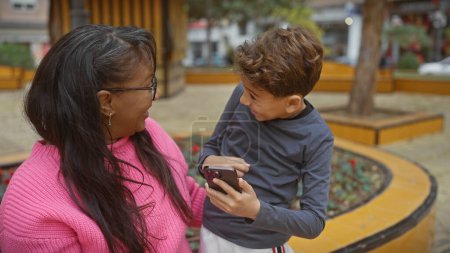 Foto de Una mujer y un niño con un smartphone disfrutando de un tiempo de calidad juntos en un colorido entorno de parque urbano. - Imagen libre de derechos