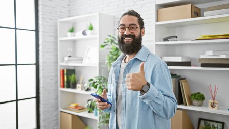 Téléchargez les photos : Homme hispanique souriant avec barbe en vêtements décontractés à l'aide d'un smartphone donne pouces vers le haut dans un cadre de bureau moderne. - en image libre de droit