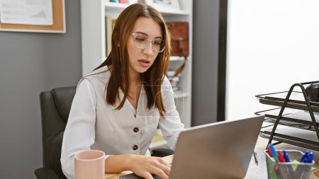 Téléchargez les photos : Une jeune femme concentrée travaillant sur un ordinateur portable dans un environnement de bureau moderne, illustrant le professionnalisme et la productivité. - en image libre de droit