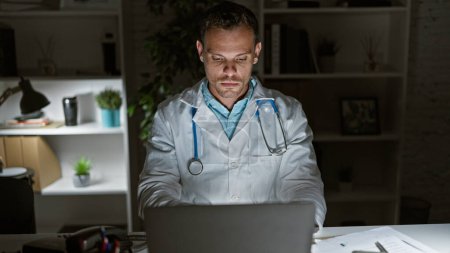 Hispanischer Arzt im Laborkittel arbeitet nachts im Klinikbüro am Laptop und ruft Hingabe hervor.