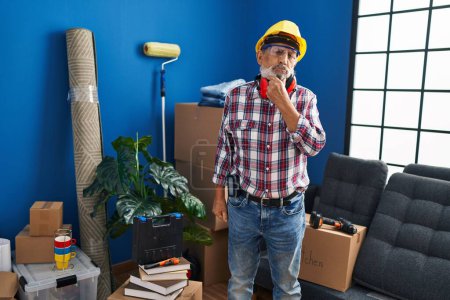Photo for Cheerful senior man in hardhat and safety glasses, confidently smiling at the camera, hand on chin, arms crossed, positively pondering in his new home - Royalty Free Image