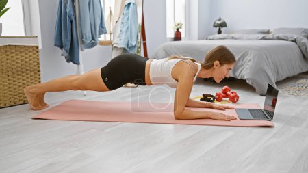 Photo for A young caucasian woman exercises in a plank position at home, following a workout video on her laptop in a spacious bedroom. - Royalty Free Image