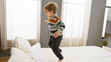 Foto de Niño rubio riendo mientras salta sobre una cama blanca en un dormitorio luminoso y soleado - Imagen libre de derechos