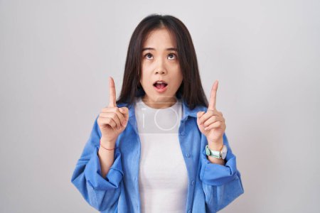 Téléchargez les photos : Jeune femme chinoise debout sur fond blanc étonné et surpris regardant vers le haut et pointant avec les doigts et les bras levés. - en image libre de droit