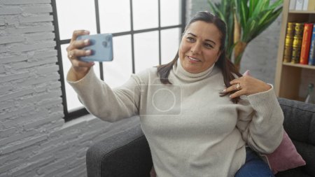Foto de Una mujer hispana sonriente se toma una selfie con un teléfono en una acogedora sala de estar, proyectando alegría y comodidad. - Imagen libre de derechos