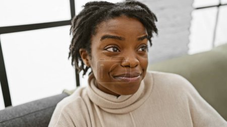 Téléchargez les photos : Une jeune femme afro-américaine souriante aux dreadlocks s'assoit avec désinvolture dans un salon intérieur, incarnant confort et authenticité. - en image libre de droit