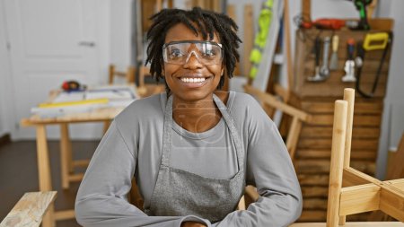 A smiling black woman with dreadlocks wearing safety glasses in a carpentry workshop.