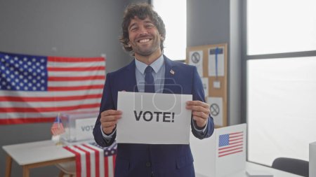 A young hispanic man with a beard holding a 'vote!' sign in a room with american flags