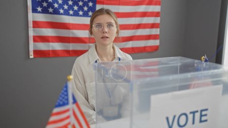 Foto de Una joven rubia con gafas está detrás de una urna electoral en una habitación con la bandera de los Estados Unidos - Imagen libre de derechos
