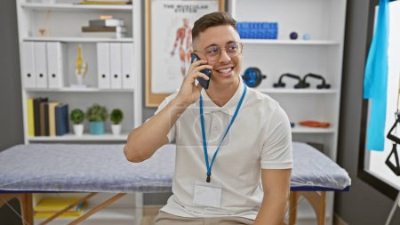 Téléchargez les photos : Un jeune hispanique souriant portant des lunettes et une lanière parle au téléphone dans une clinique de thérapie moderne. - en image libre de droit