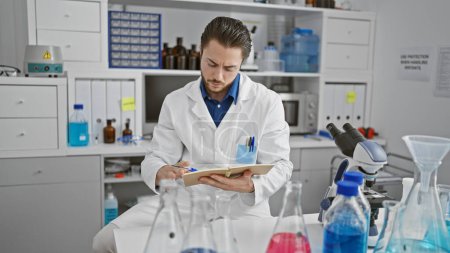 Photo for Young hispanic man scientist writing on notebook sitting at laboratory - Royalty Free Image