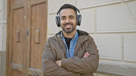 Photo for A smiling hispanic man with a beard wearing headphones outdoors in an urban city setting, exuding casual confidence. - Royalty Free Image