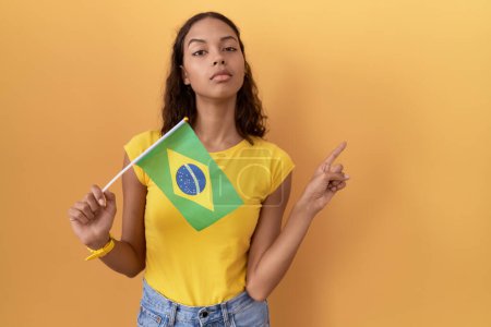 Téléchargez les photos : Jeune femme hispanique tenant drapeau brésilien pointant avec le doigt de la main sur le côté montrant la publicité, visage sérieux et calme - en image libre de droit