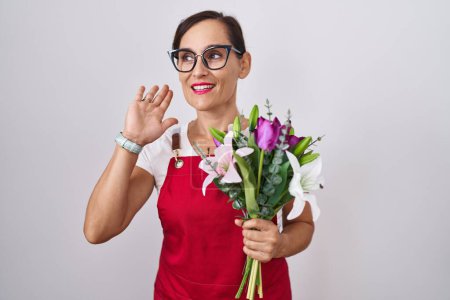 Téléchargez les photos : Femme brune d'âge moyen portant un tablier travaillant dans un magasin de fleuristes tenant un bouquet souriant avec la main sur l'oreille en écoutant une rumeur ou des commérages. concept de surdité. - en image libre de droit