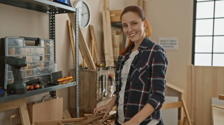 Foto de Una joven sonriente con el pelo rojo se para en un taller de carpintería rodeado de herramientas y madera. - Imagen libre de derechos