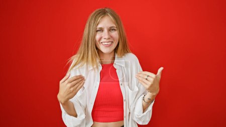 Une jeune femme blonde souriante en chemise blanche et haut rouge sur un fond rouge isolé fait des gestes conversationnels.