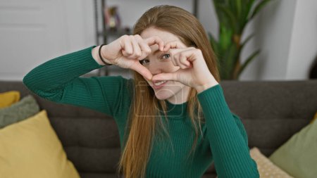 A cheerful young woman with long blonde hair, wearing a green sweater, makes a heart shape with her hands indoors.
