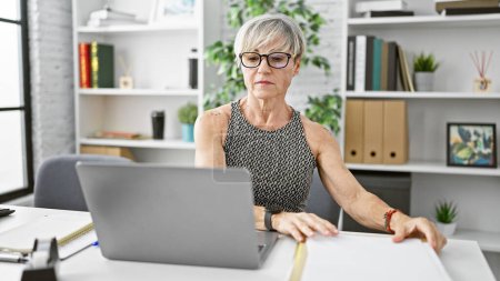 Photo for A mature woman with short grey hair works attentively at her laptop in a modern office interior. - Royalty Free Image
