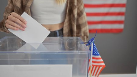 Una joven mujer caucásica votando en un colegio electoral en los EE.UU., con una bandera americana presente en la sala.