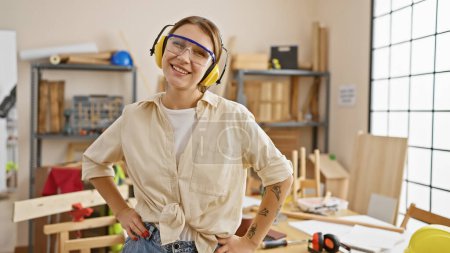 Photo for Smiling woman with safety goggles and headphones standing confidently in a sunny carpentry workshop. - Royalty Free Image