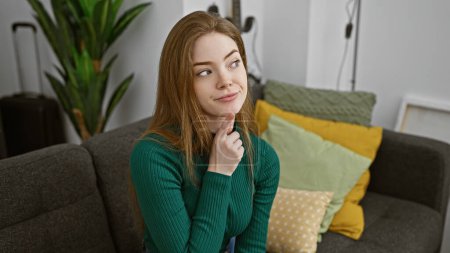 Foto de Una joven reflexiva se sienta en un sofá en una acogedora sala de estar, contemplando con una maleta cerca, sugiriendo viajar. - Imagen libre de derechos